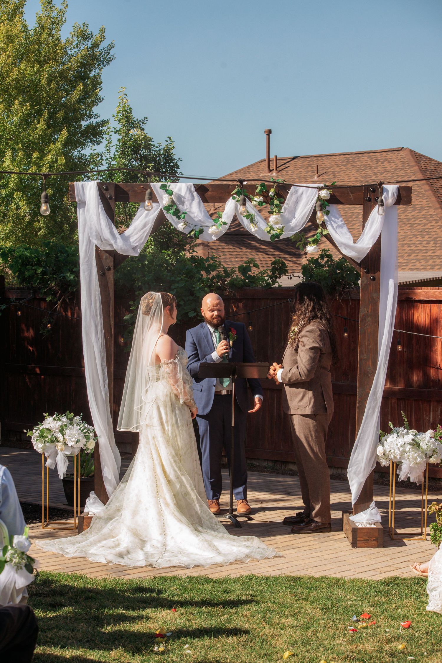 Bride and groom exchange vows under a decorated wooden arch with white draping and greenery at an outdoor wedding.