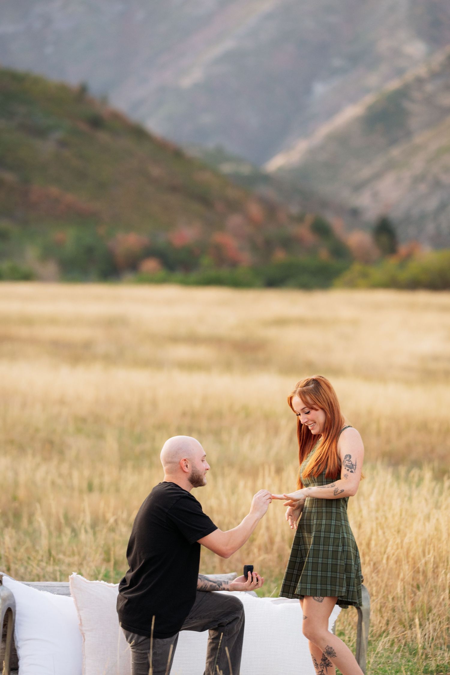 Bald tattooed man pointing at redhead woman showing off engagement ring outdoors with mountain backdrop.