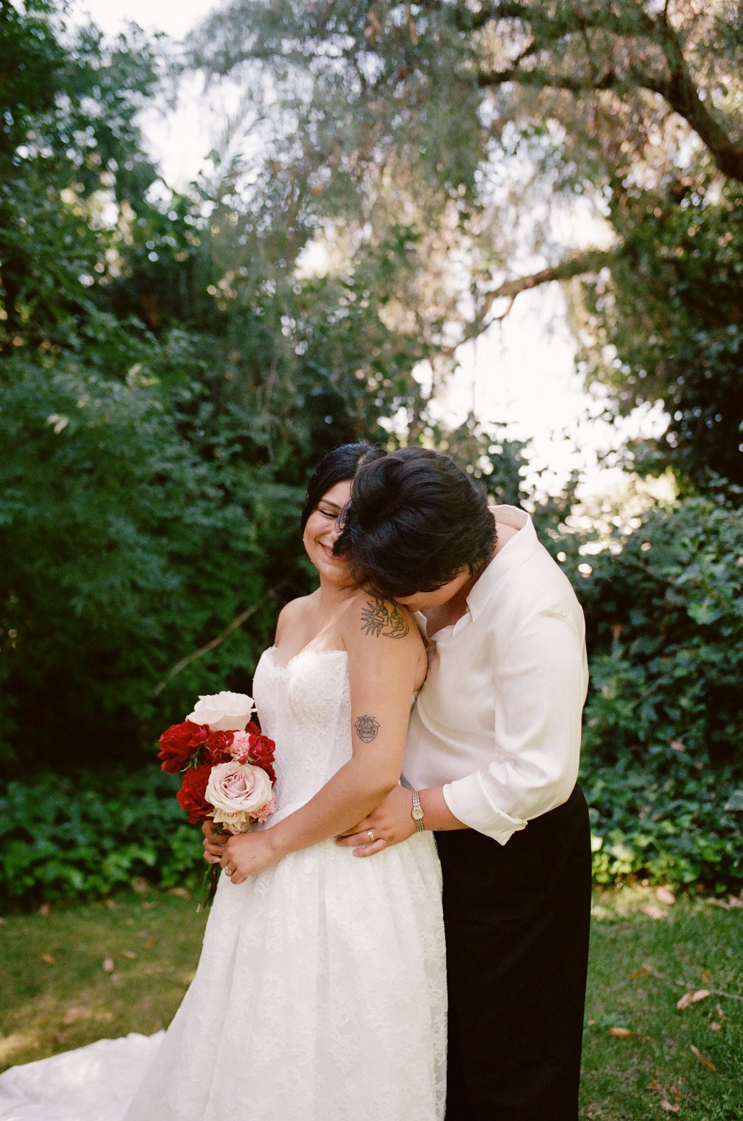 Bride in a white lace wedding dress holding a red and pink bouquet embraces her partner in a white shirt in a leafy garden.