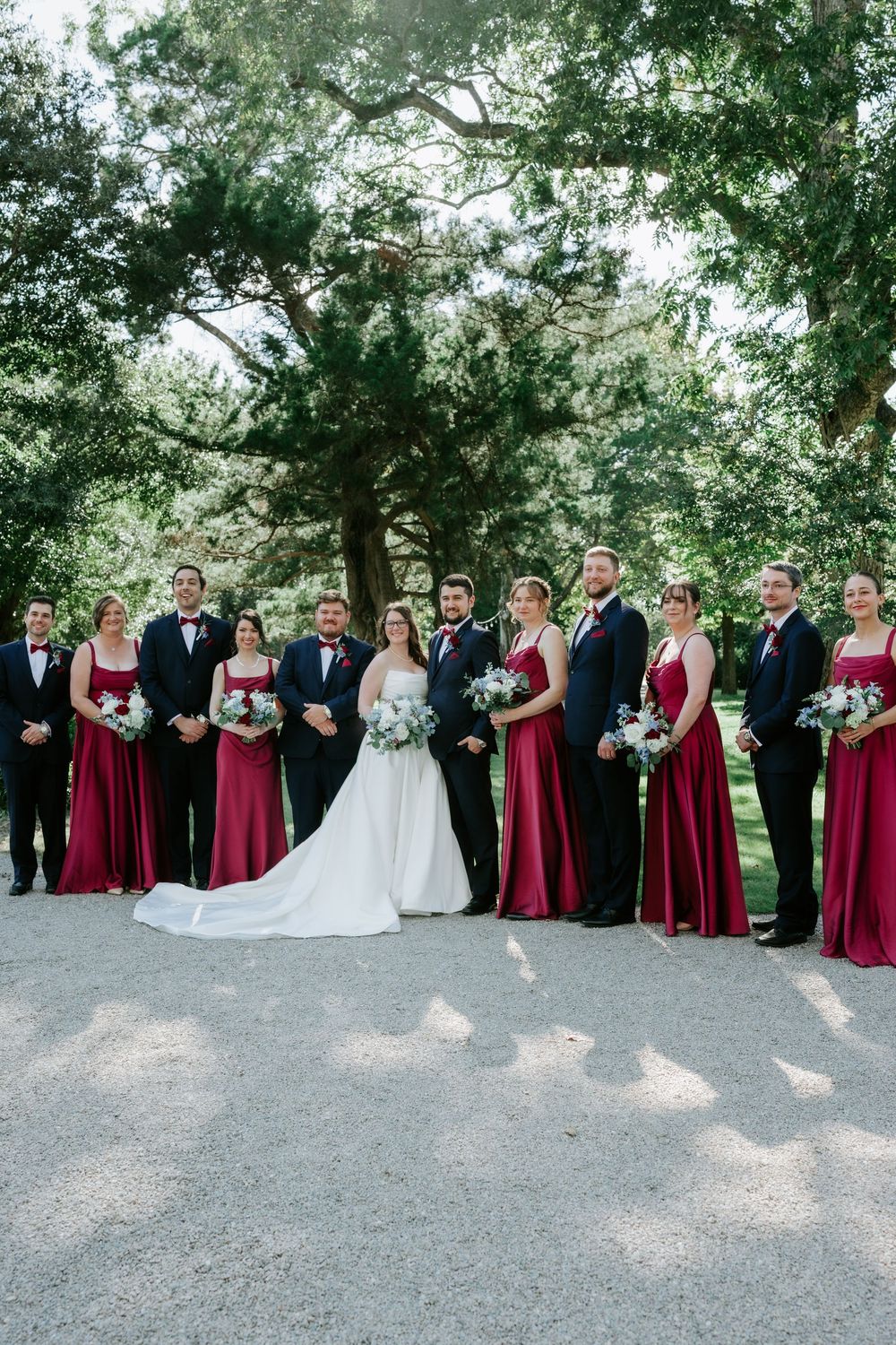 Wedding party poses outdoors, bride in white gown flanked by groomsmen in navy suits and bridesmaids in burgundy dresses.