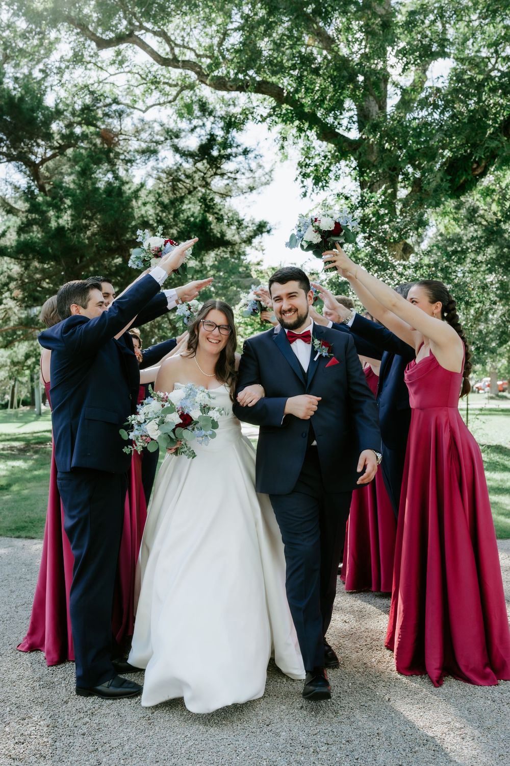Wedding party walks outdoors, bride in white gown with bouquet, groomsmen in navy suits, bridesmaids in burgundy dresses raising glasses.