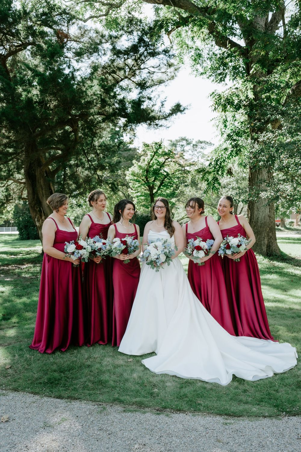 Bride in white gown with five bridesmaids in burgundy dresses holding bouquets outdoors under large trees.