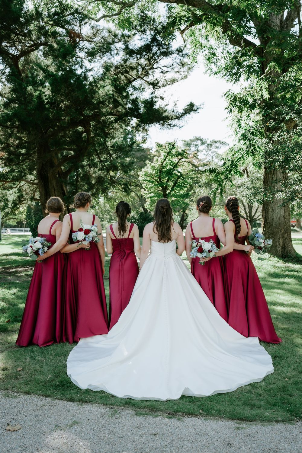 Bride in white gown with four bridesmaids in red dresses holding bouquets, viewed from behind in a lush green park.