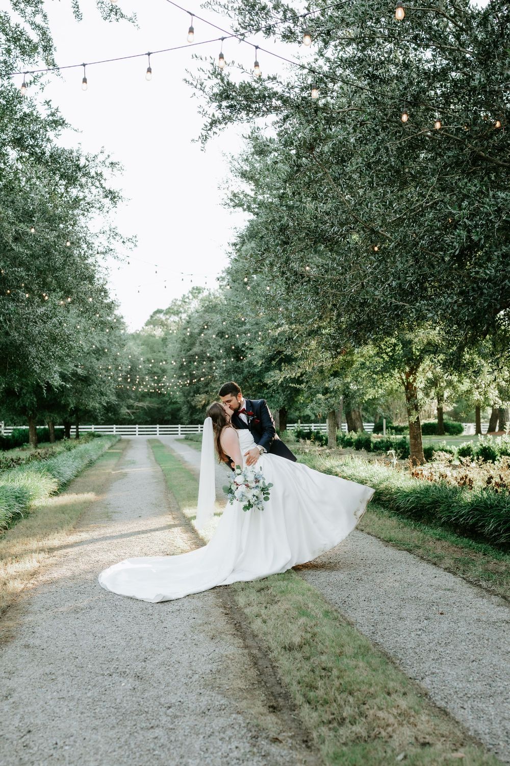 Bride and groom share a romantic dip kiss on a tree-lined path with string lights at an outdoor wedding venue.