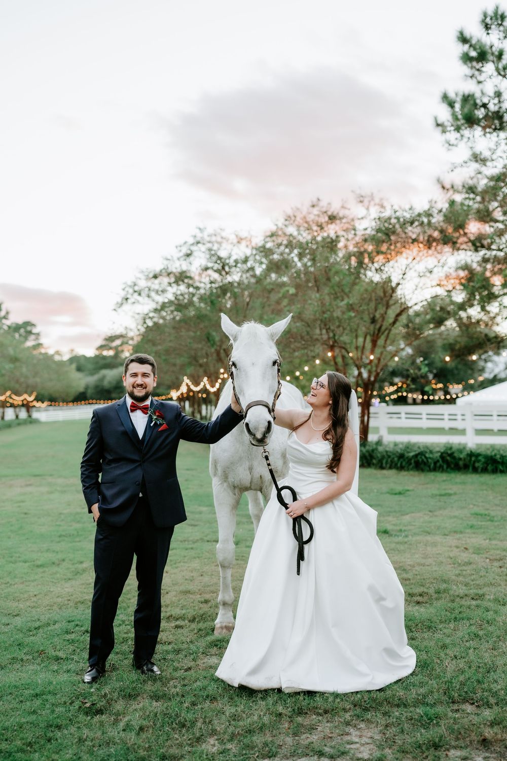 Bride and groom posing with a white horse on a green lawn at sunset during their outdoor wedding celebration.