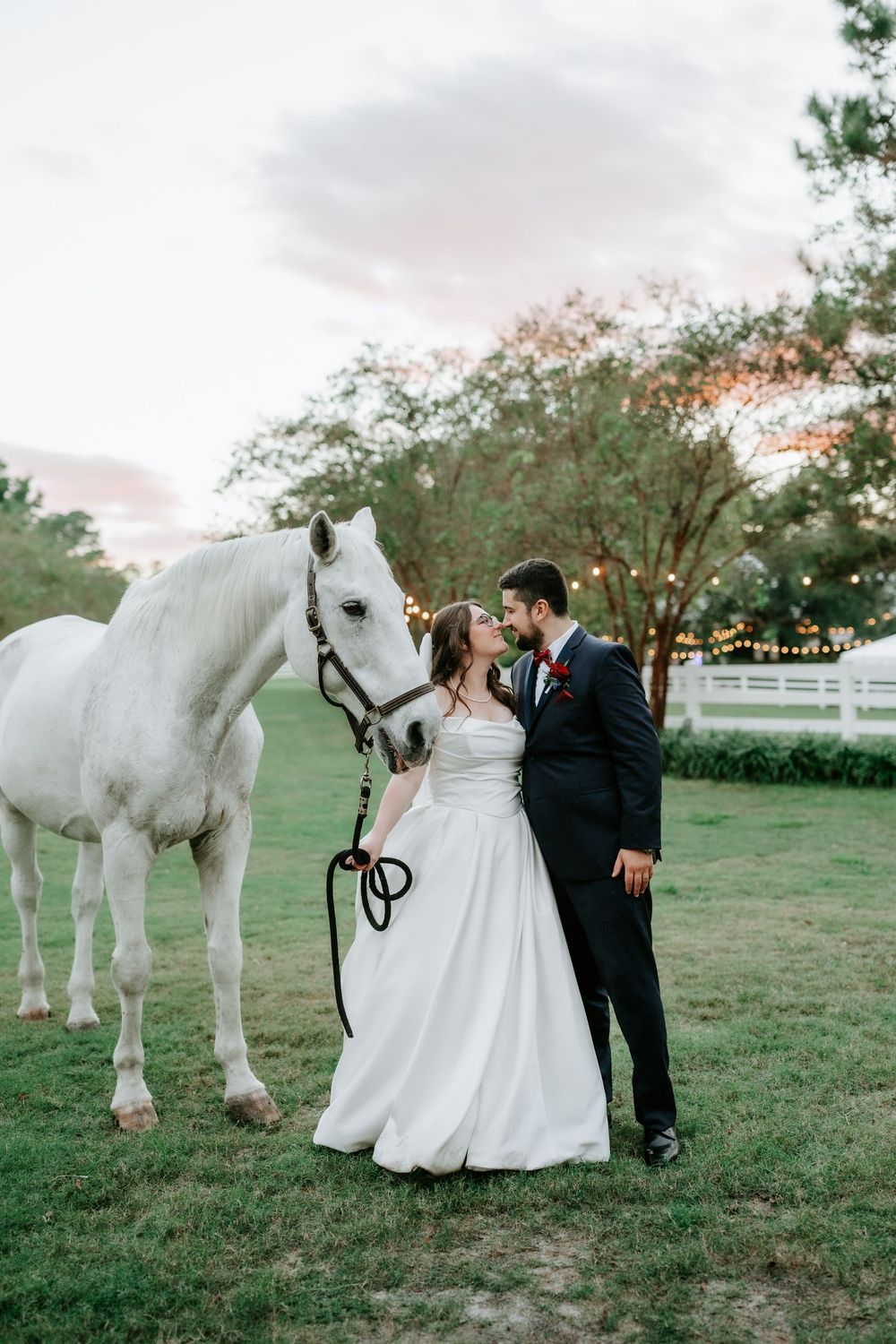 Bride and groom share a romantic moment beside a white horse at sunset on a green lawn with string lights.