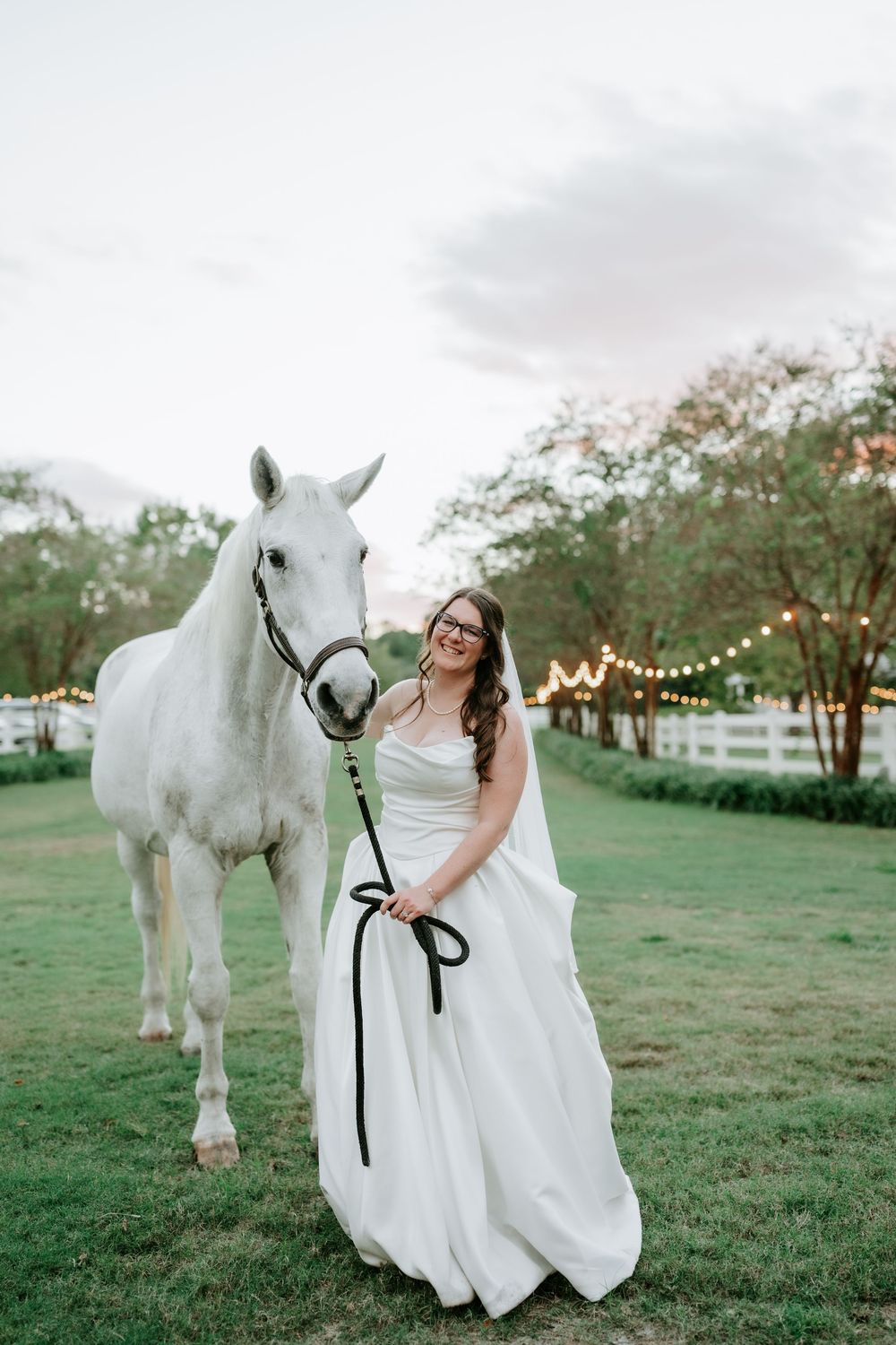 Bride in white gown holding a white horse on a lush green lawn with string lights at dusk.