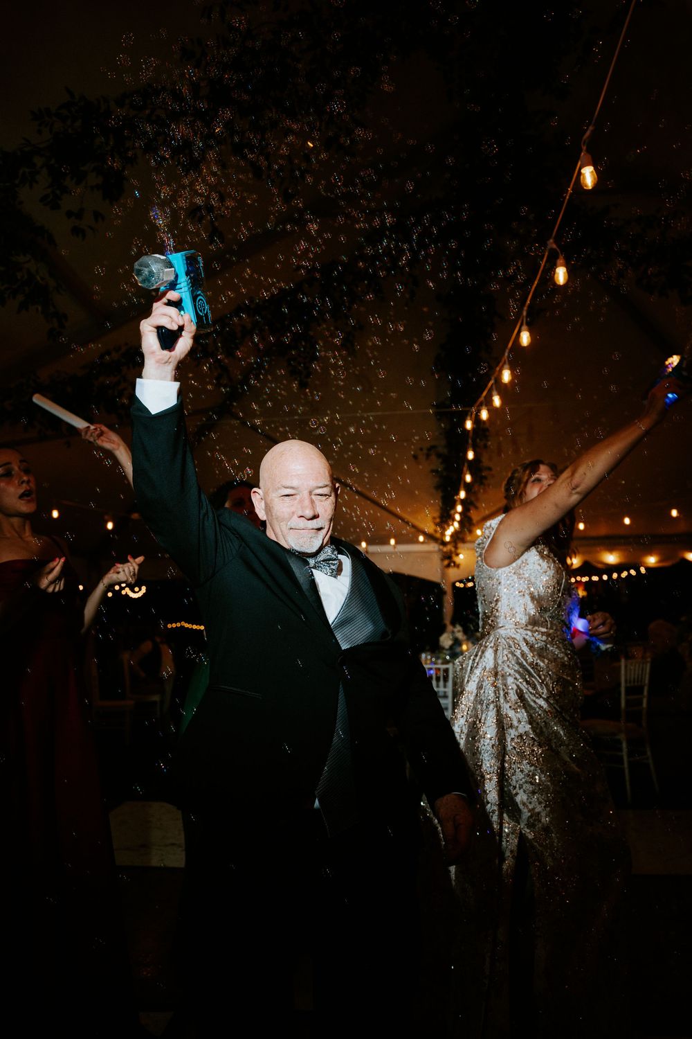 Elderly man in tuxedo celebrating at wedding reception, raising drink amid confetti under string lights.