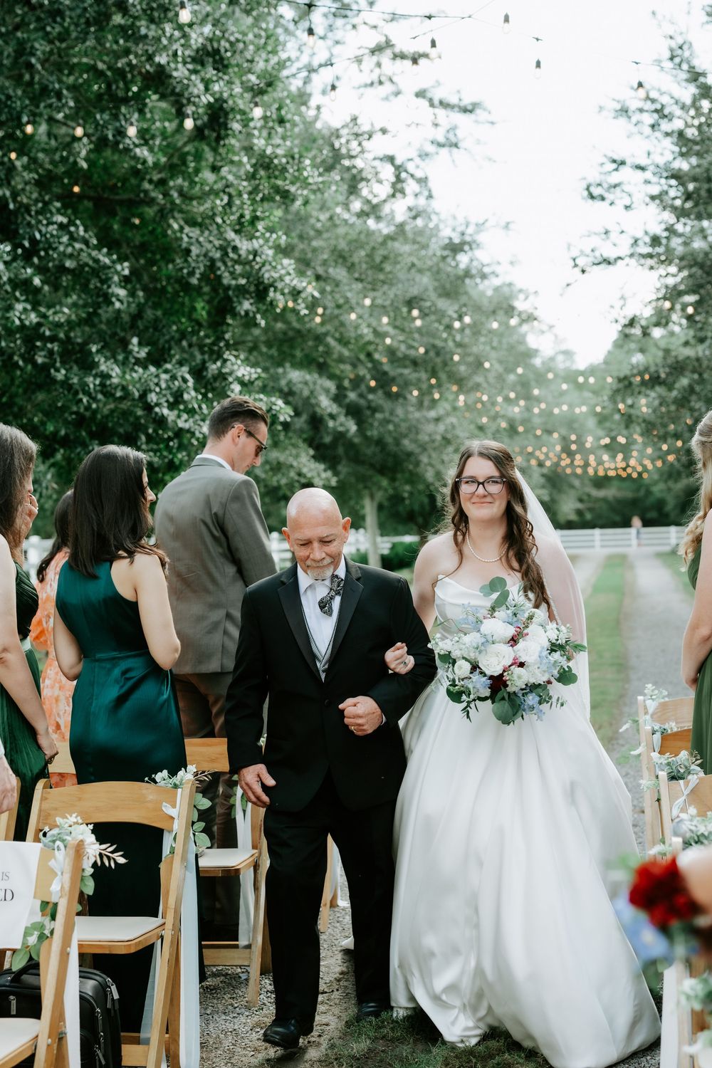 Smiling bride walks down the outdoor aisle with her father during a garden wedding ceremony with string lights.