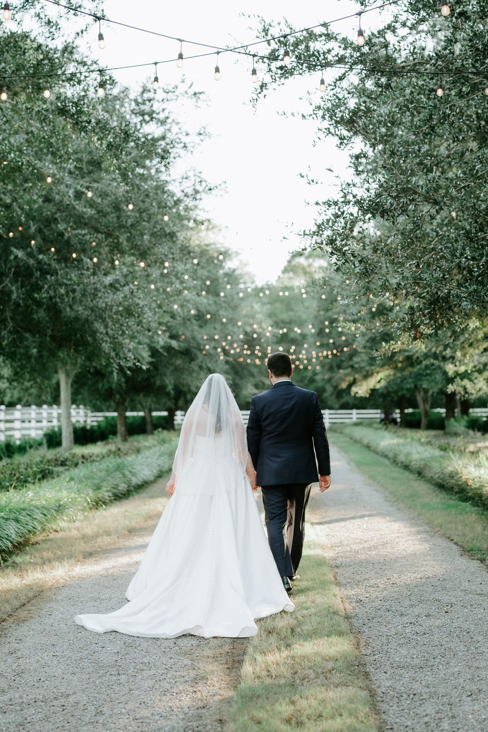 Bride and groom walking hand in hand down a tree-lined path with string lights at an outdoor wedding venue.