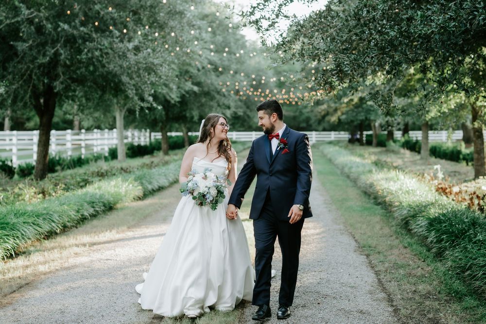 Bride and groom walk hand-in-hand down a tree-lined path with string lights at an elegant outdoor wedding venue.