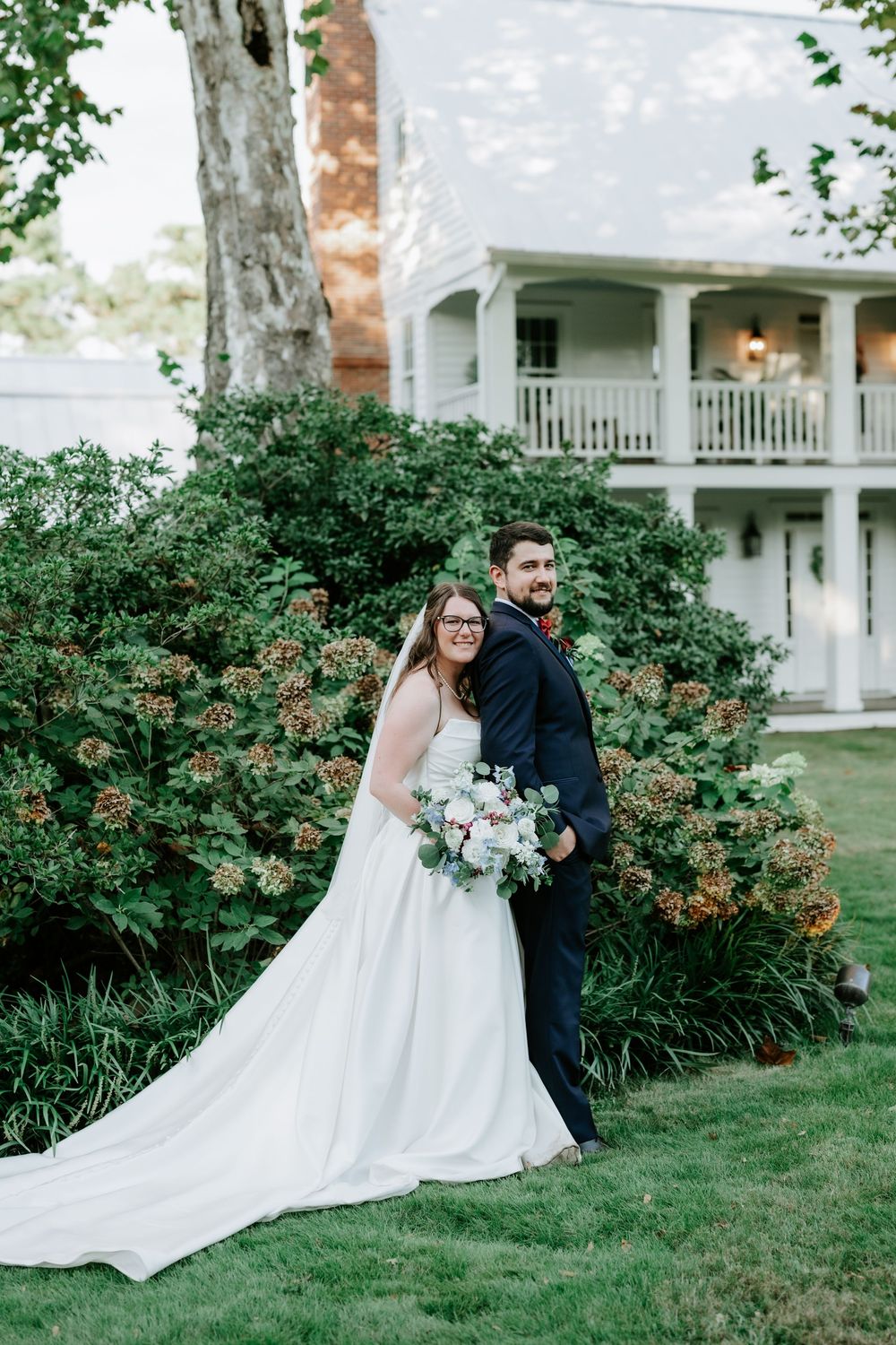 Bride and groom pose back-to-back in front of lush greenery and a white colonial house on their wedding day.