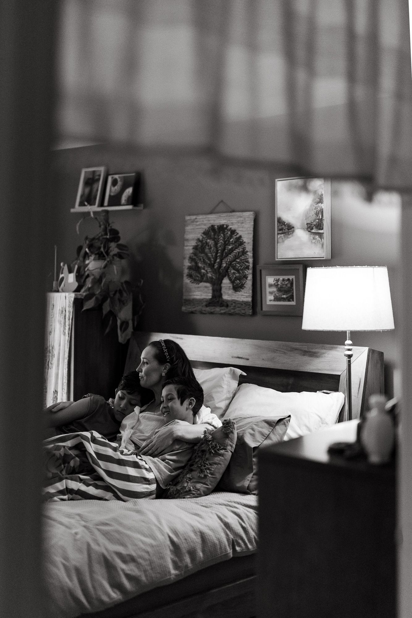 Black and white photo of two people cuddling on a bed in a cozy bedroom with framed art and a glowing lamp.