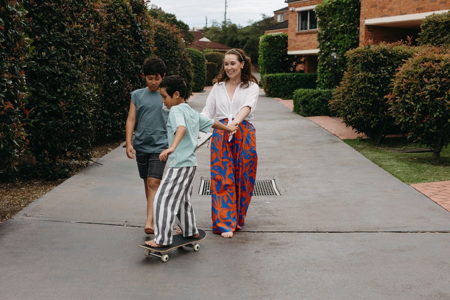 Woman in colorful pants walks with two boys on a skateboard down a suburban driveway lined with hedges.