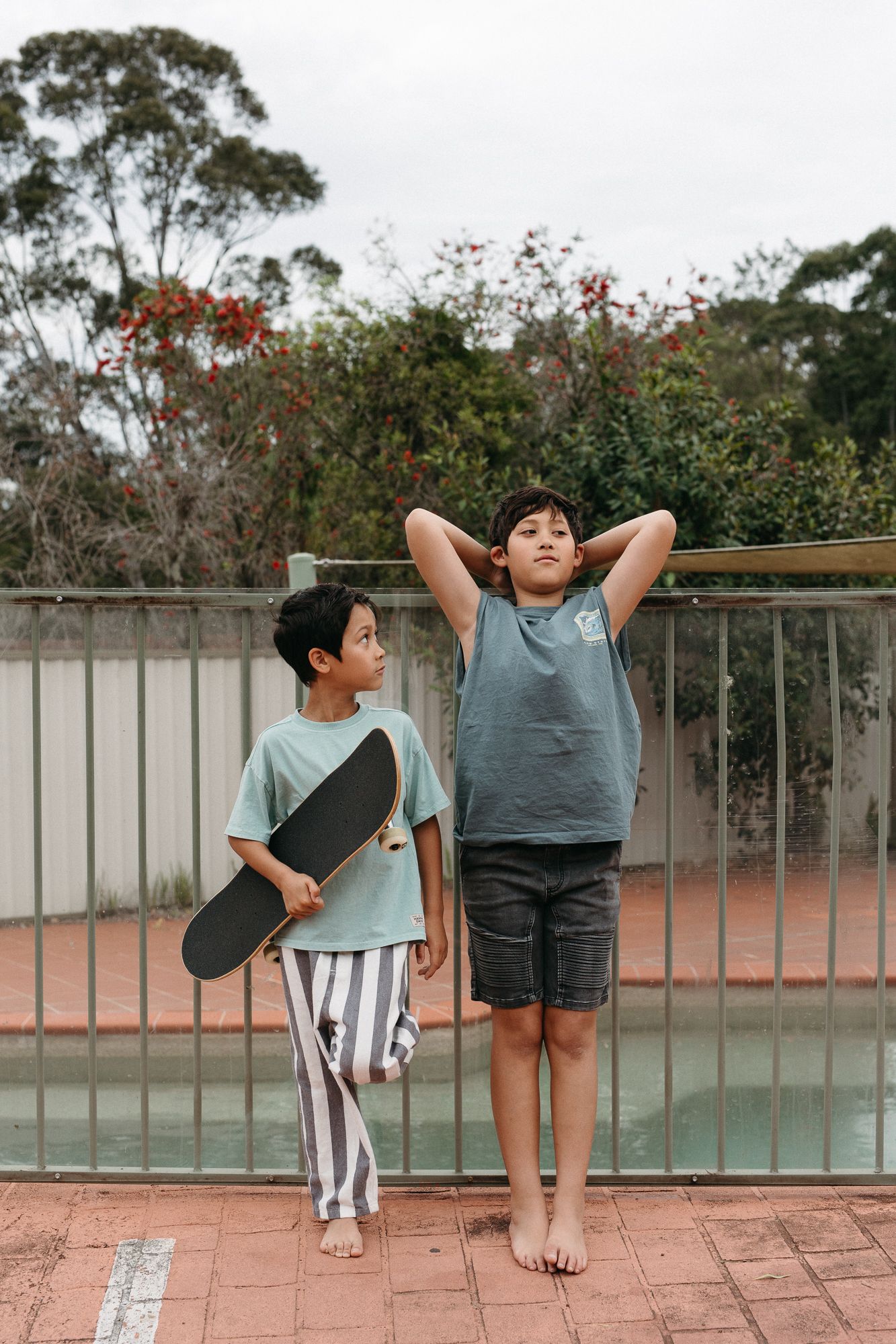 Two boys stand by a pool fence outdoors, one holding a skateboard in striped pants, the other in a gray tank top.