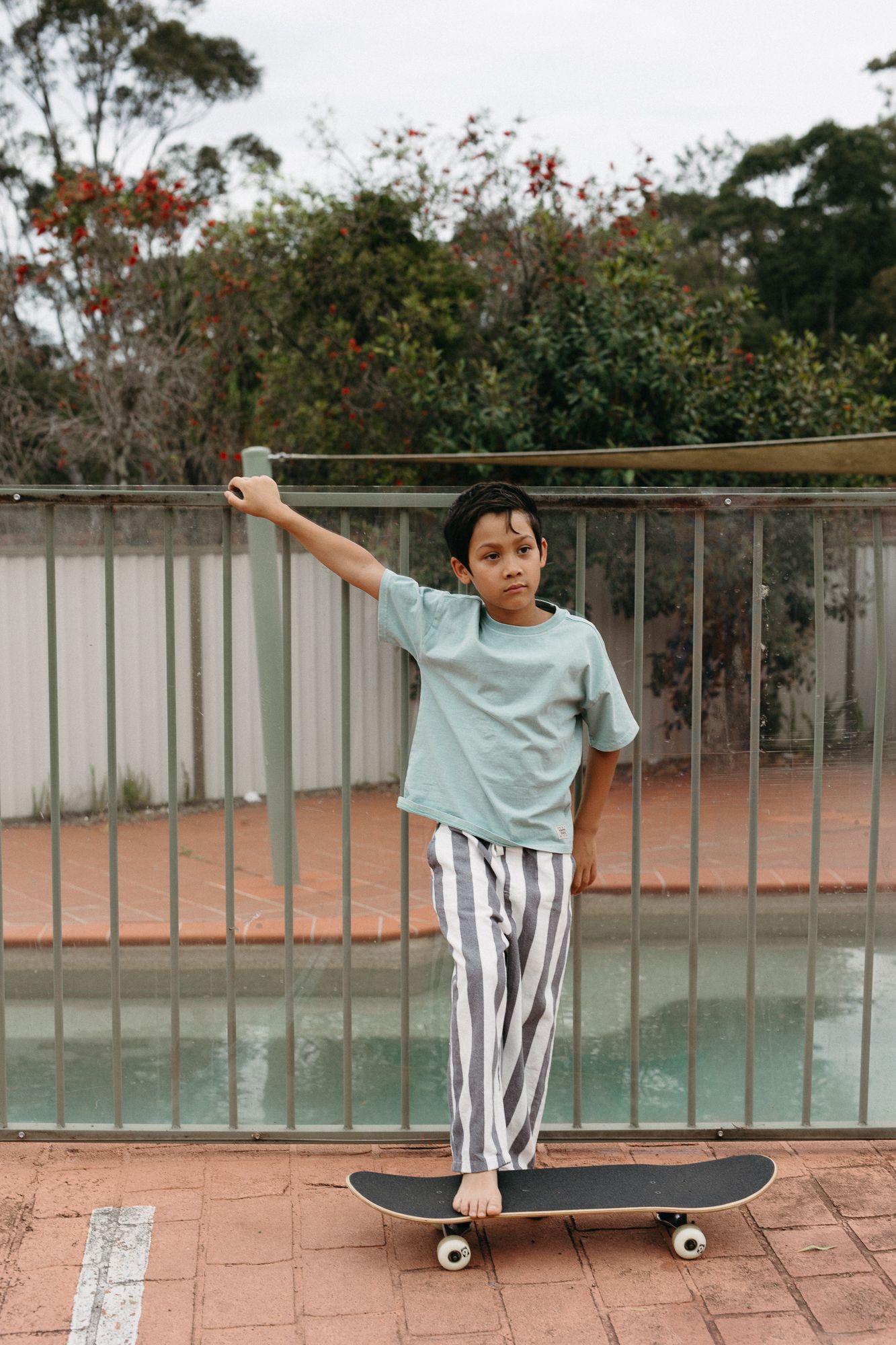 A young boy in a mint green shirt and striped pants stands on a skateboard holding a railing outdoors.