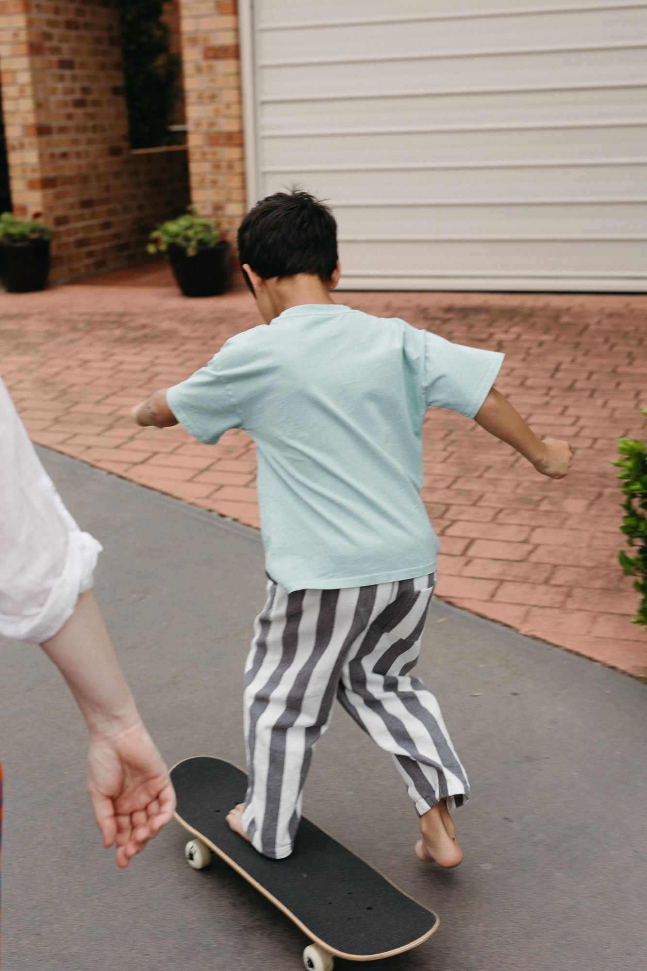 Young boy in teal shirt and striped pants learning to skateboard on driveway, held by adult hand.