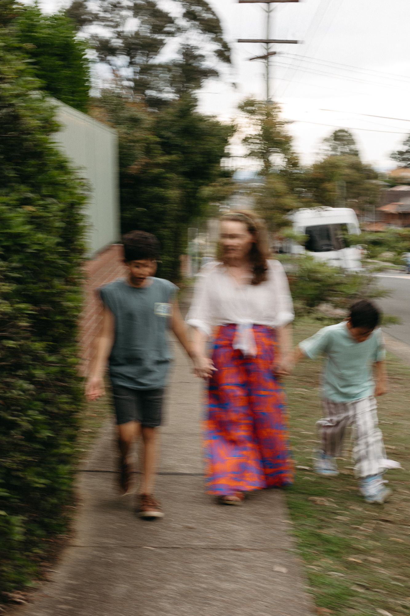 A woman in a colorful maxi skirt walks hand-in-hand with two children on a suburban sidewalk.