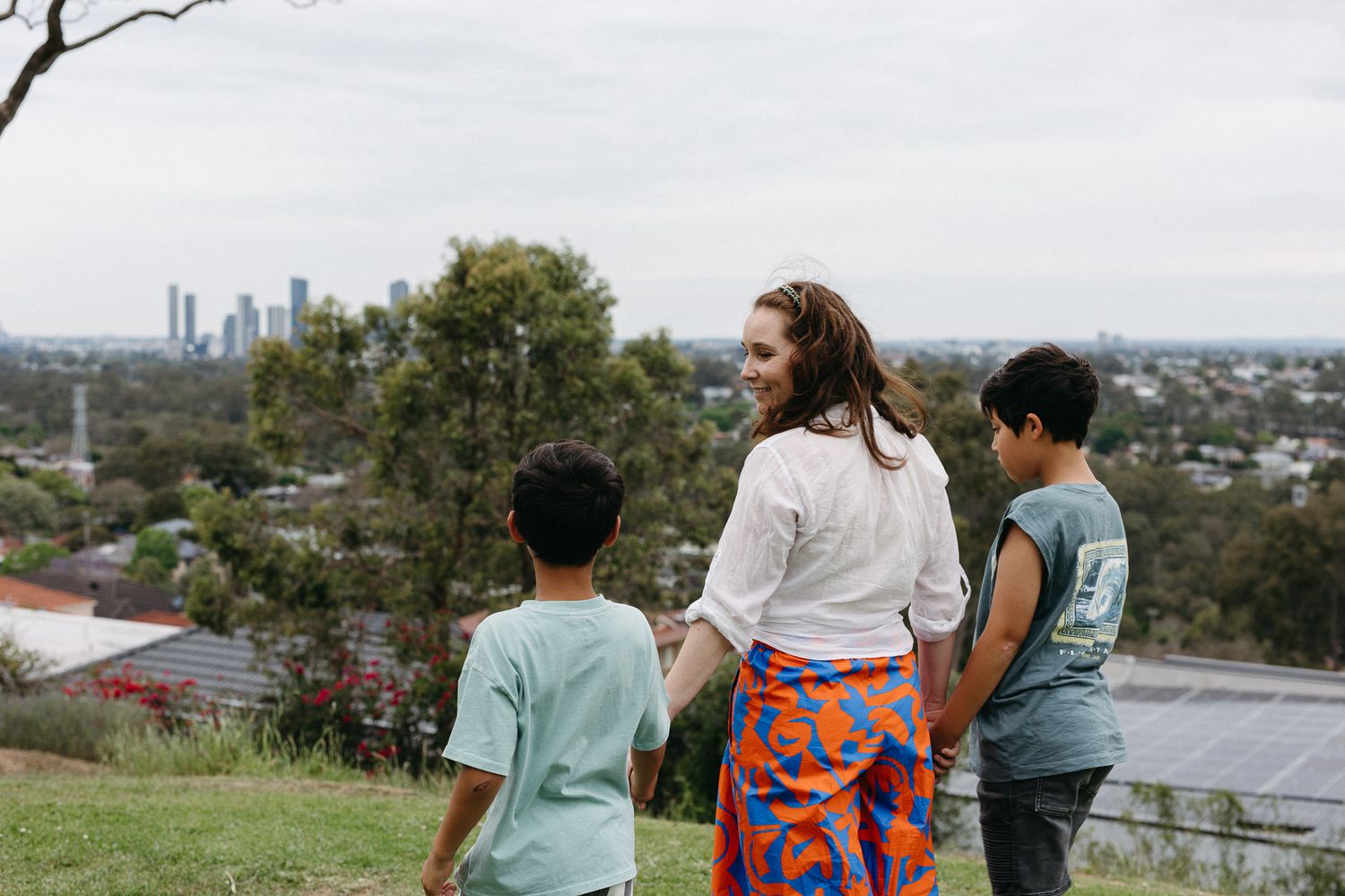 Woman and two children stand on a grassy hill overlooking a city skyline surrounded by trees.
