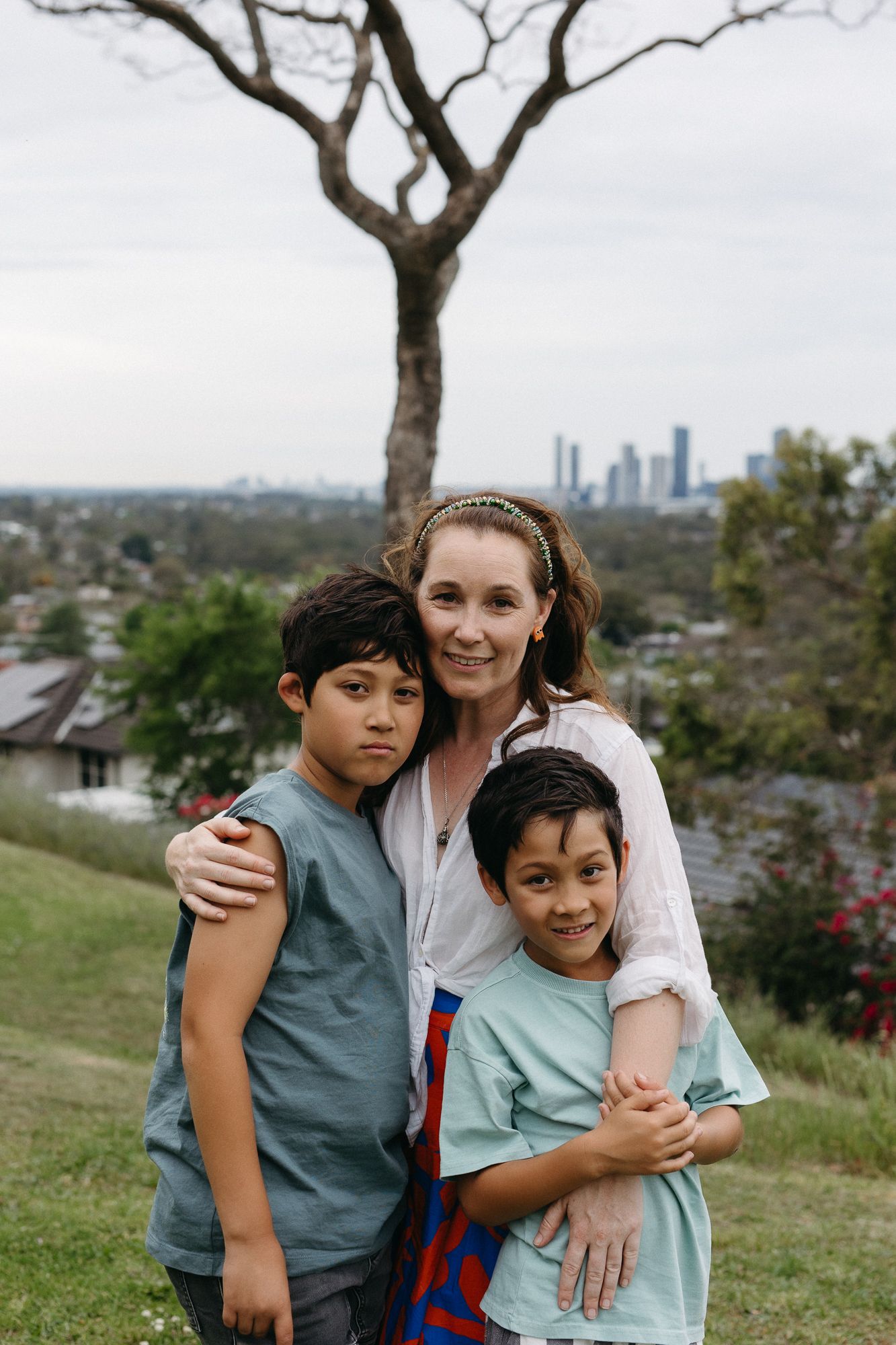 A woman smiles while hugging two young boys on a grassy hill with a city skyline in the background.
