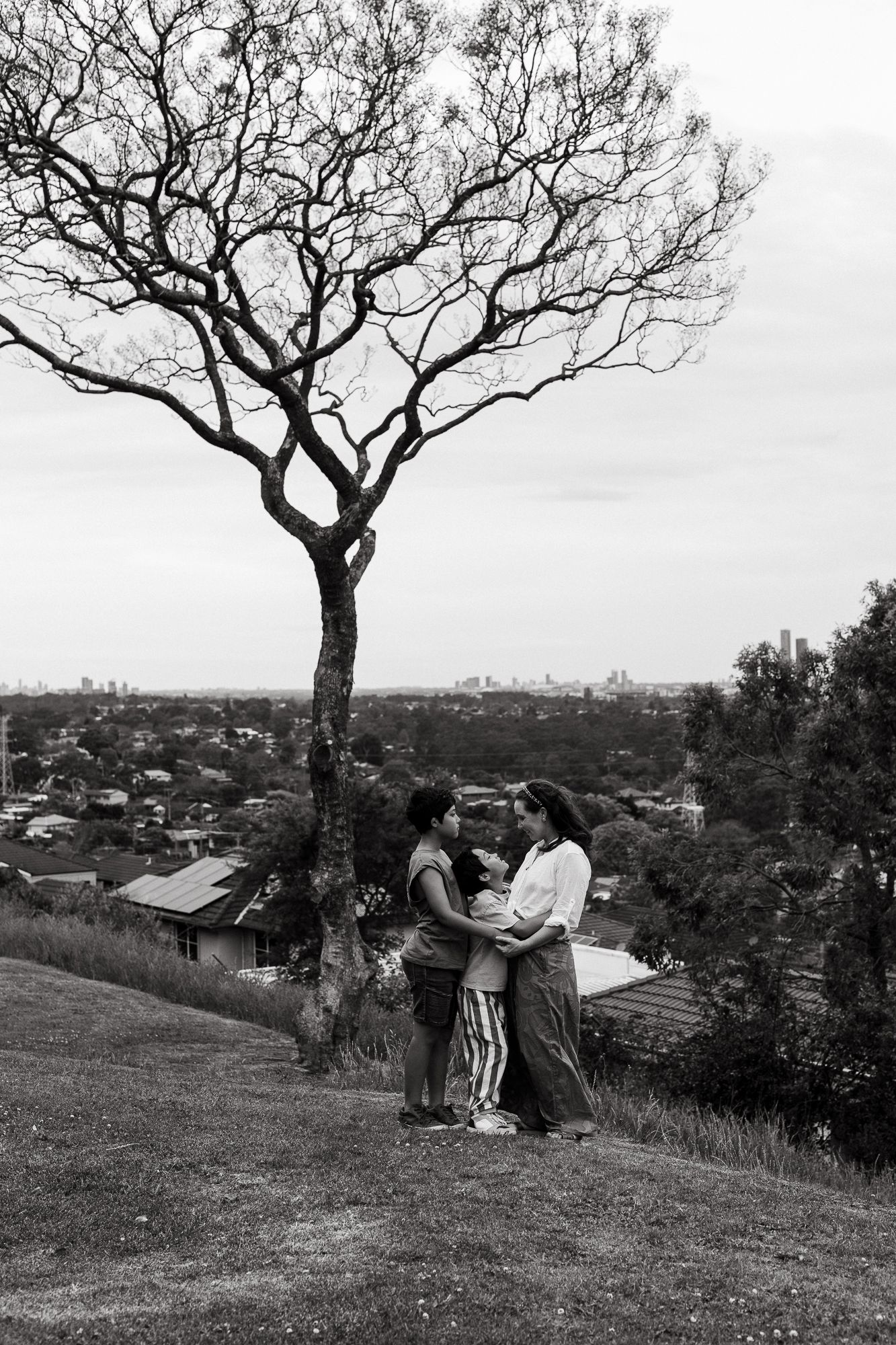 Black and white photo of a family embracing on a hill under a bare tree with a city skyline in the background.