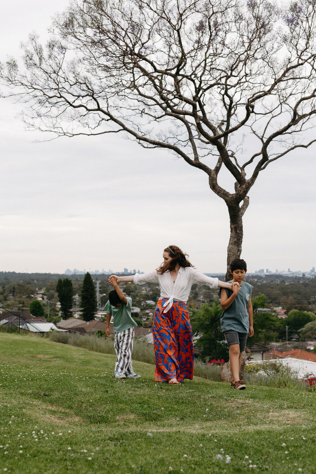 A woman holds hands with two children on a grassy hill, with a bare tree and city skyline in the background.