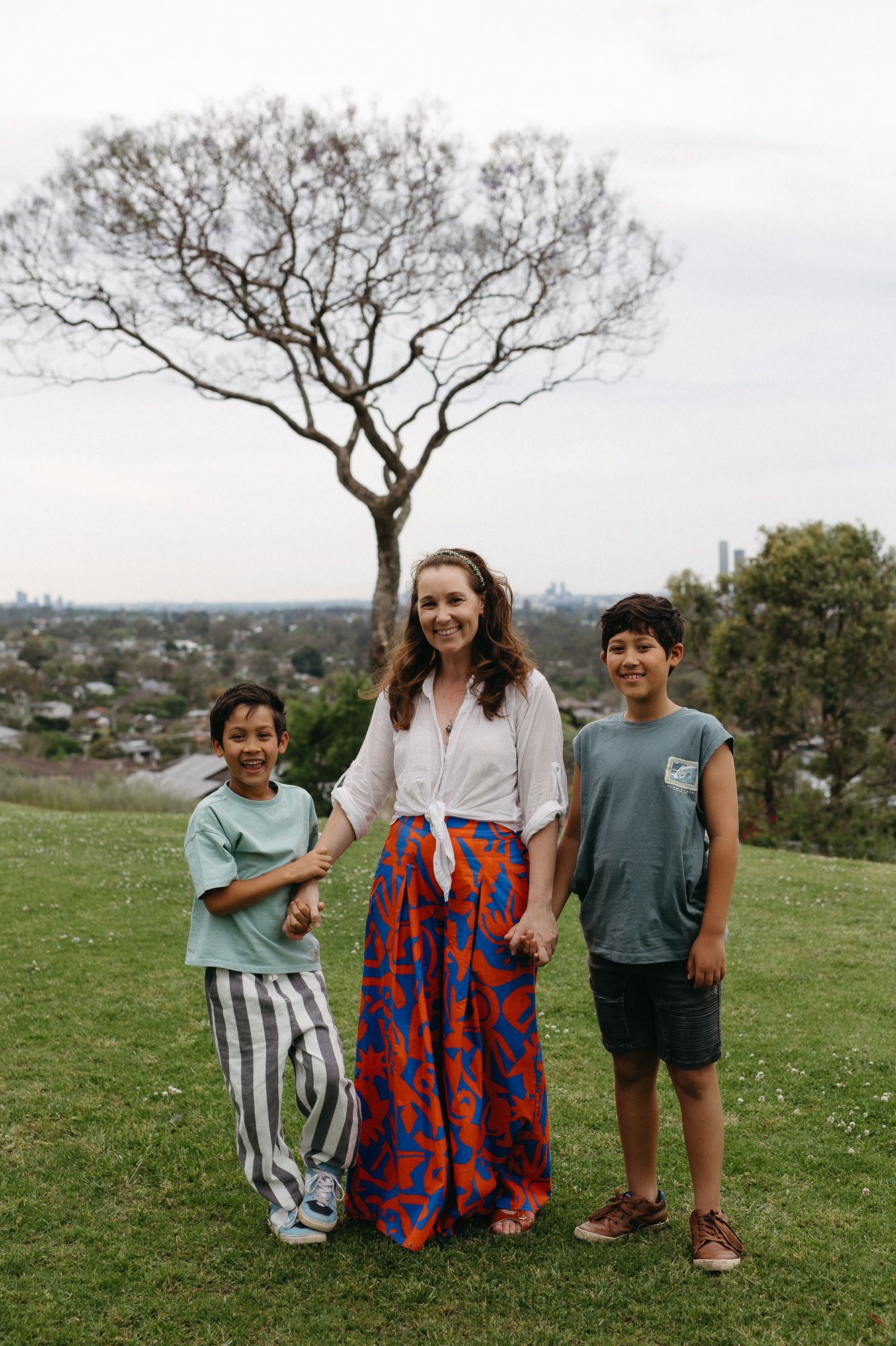 Woman in colorful maxi skirt stands with two children on grassy hill with city skyline and bare tree backdrop.
