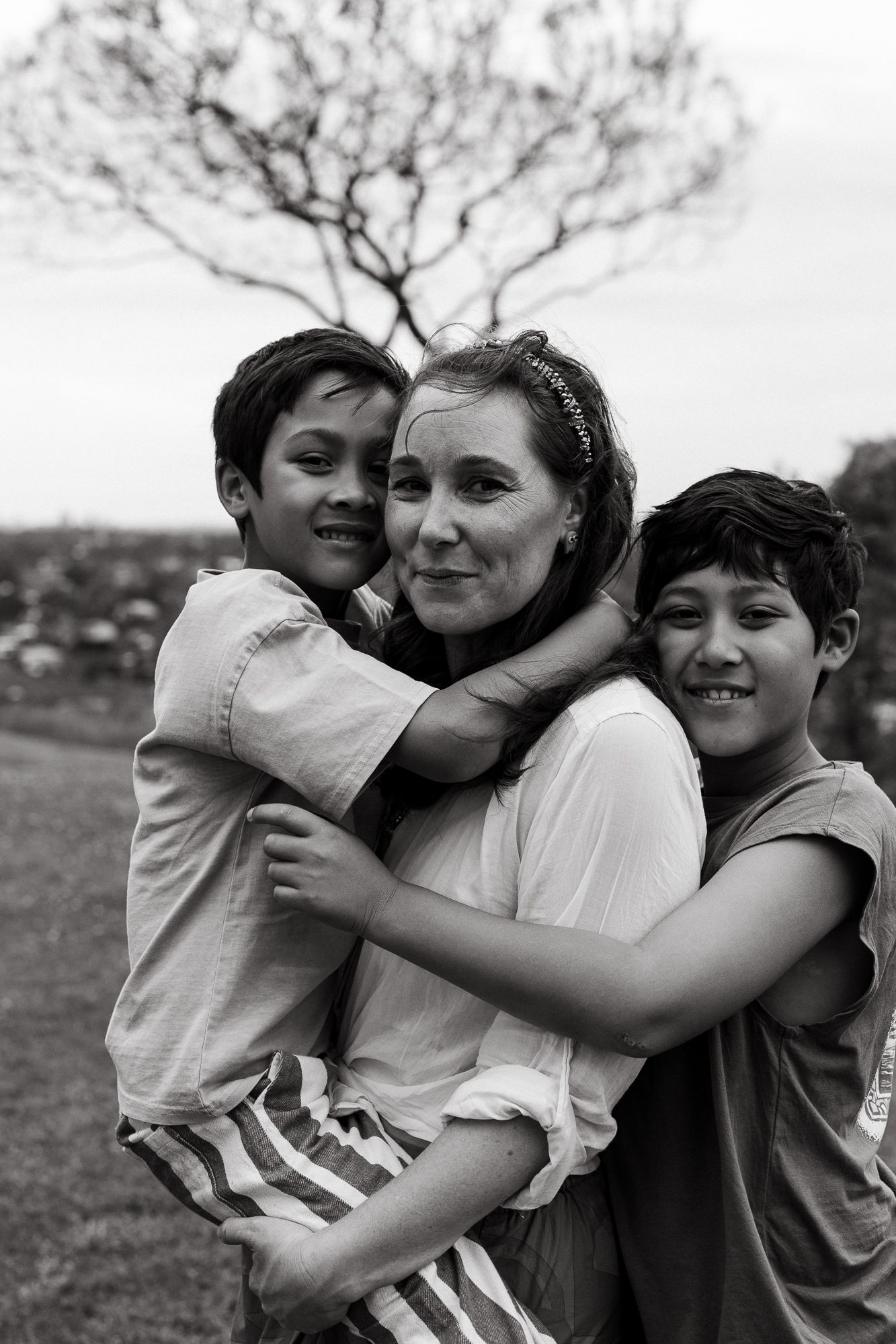 Black and white photo of a woman hugging two young boys outdoors, smiling warmly with a tree in the background.
