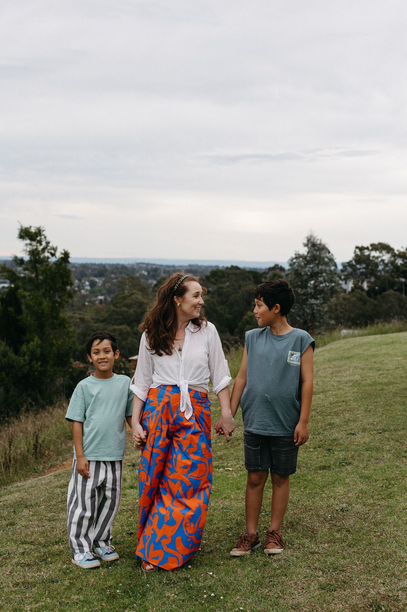 A woman in a colorful skirt stands smiling with two boys on a grassy hill with trees in the background.