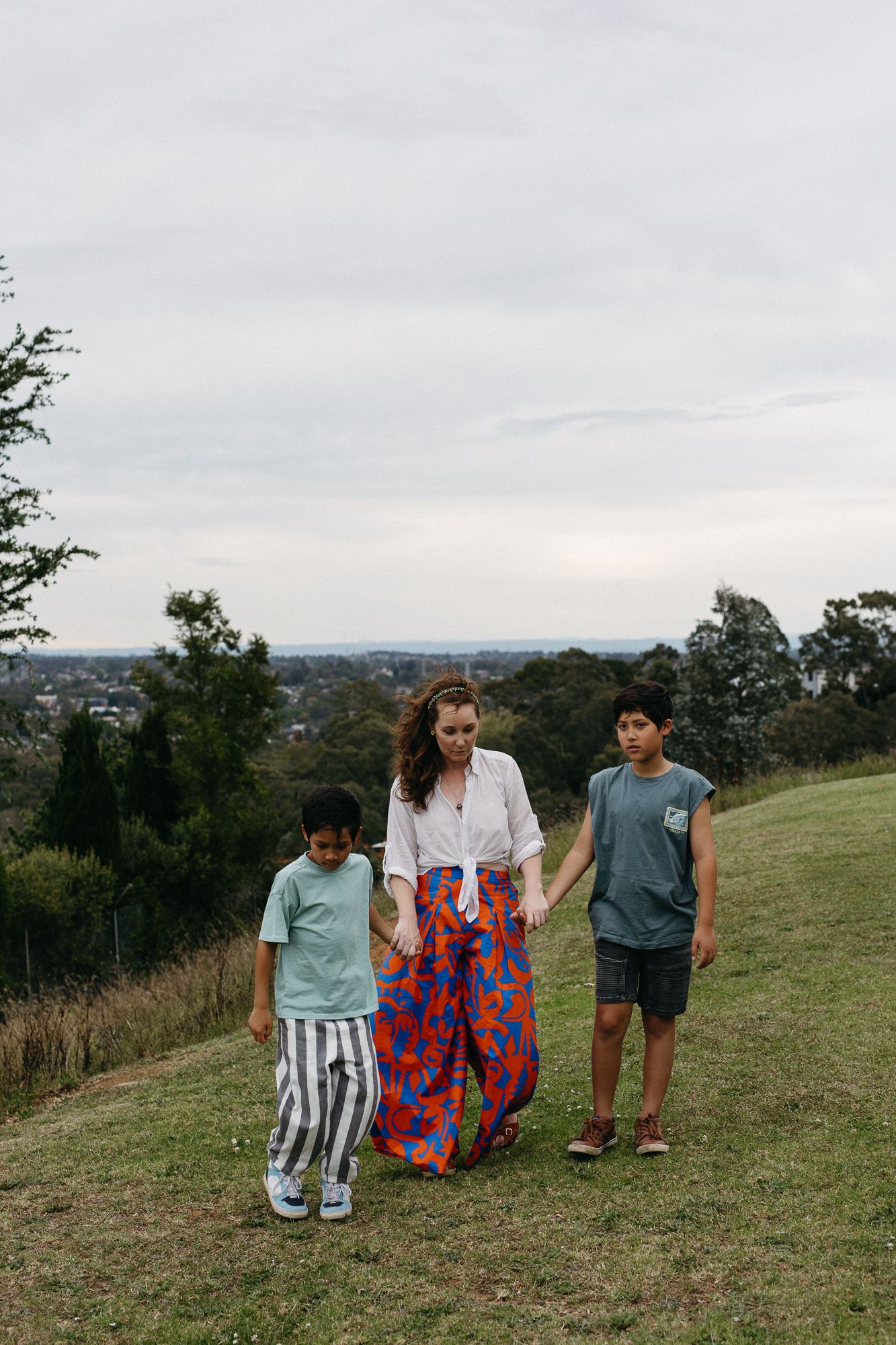 A woman in a floral skirt walks hand-in-hand with two boys on a grassy hill with a town view.