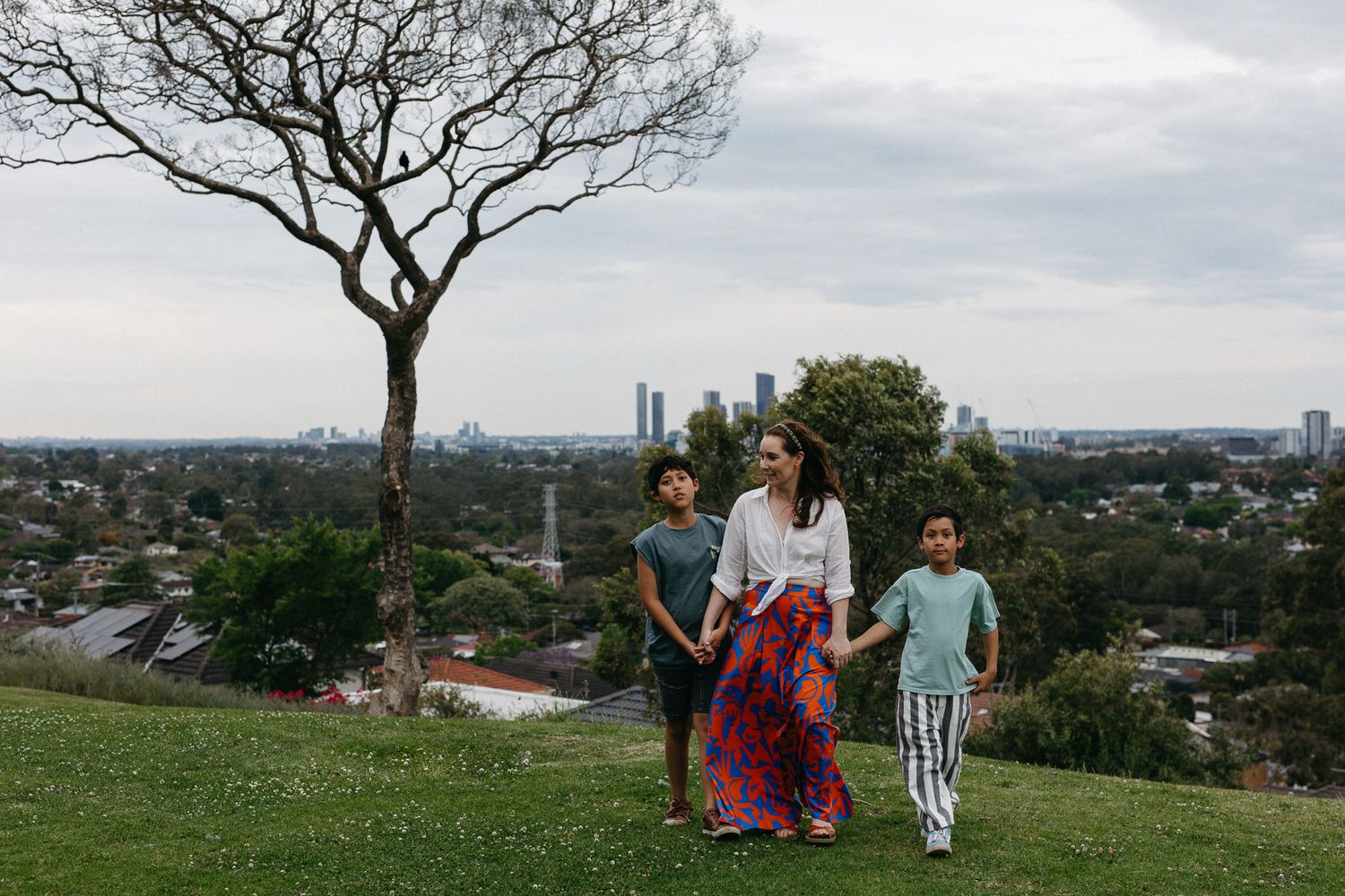 A woman and two children walk on a grassy hill with a city skyline and bare tree in the background.