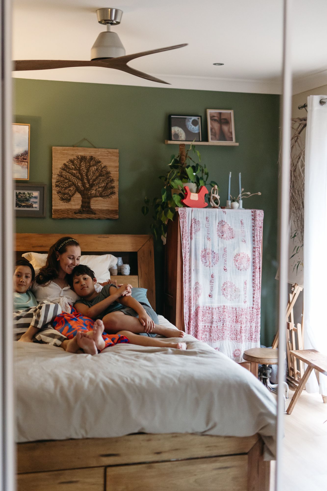 A mother cuddles with her two children on a bed in a cozy green bedroom.