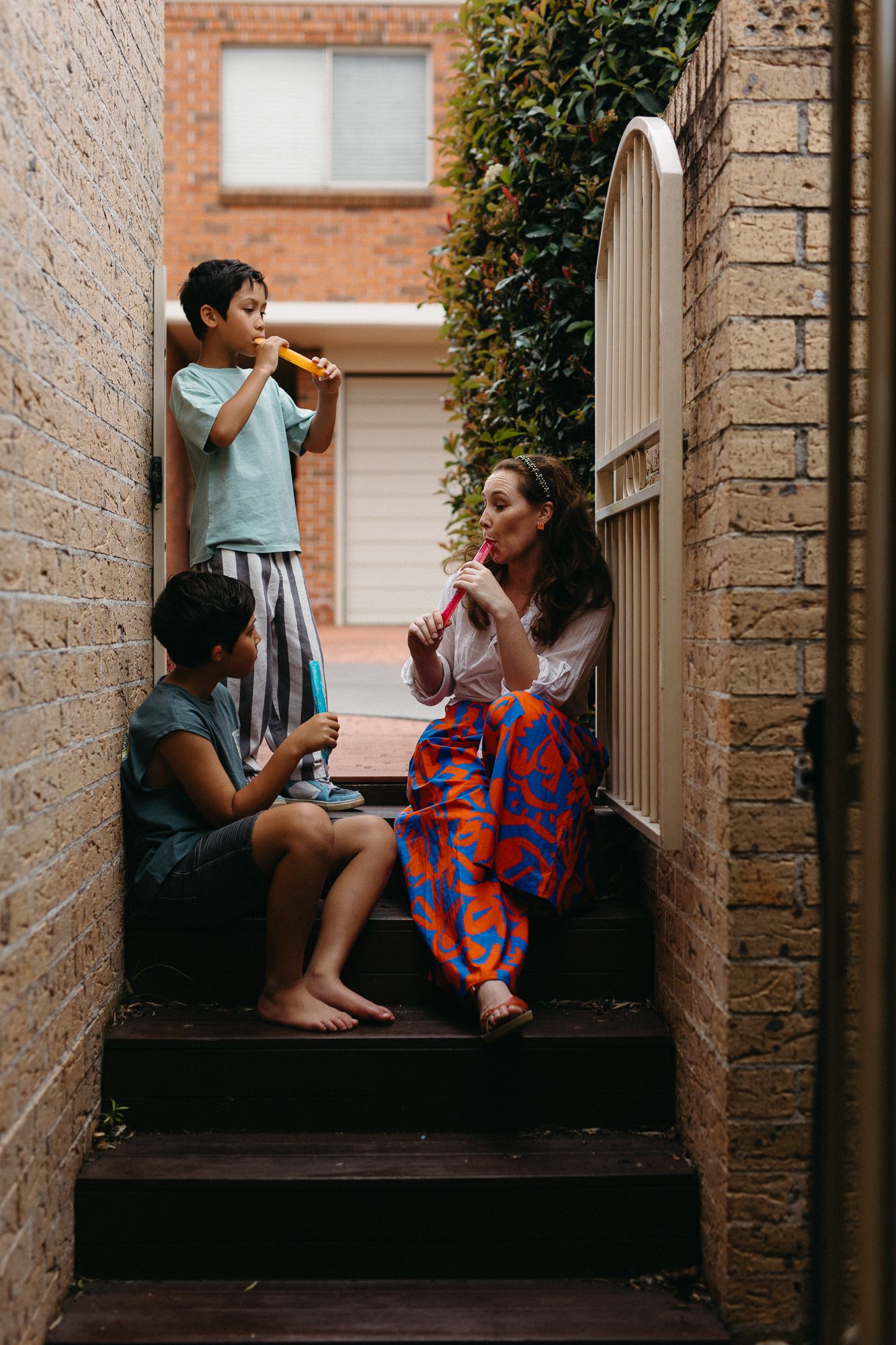 A woman and two children sit on outdoor steps eating popsicles beside a brick wall and green hedge.