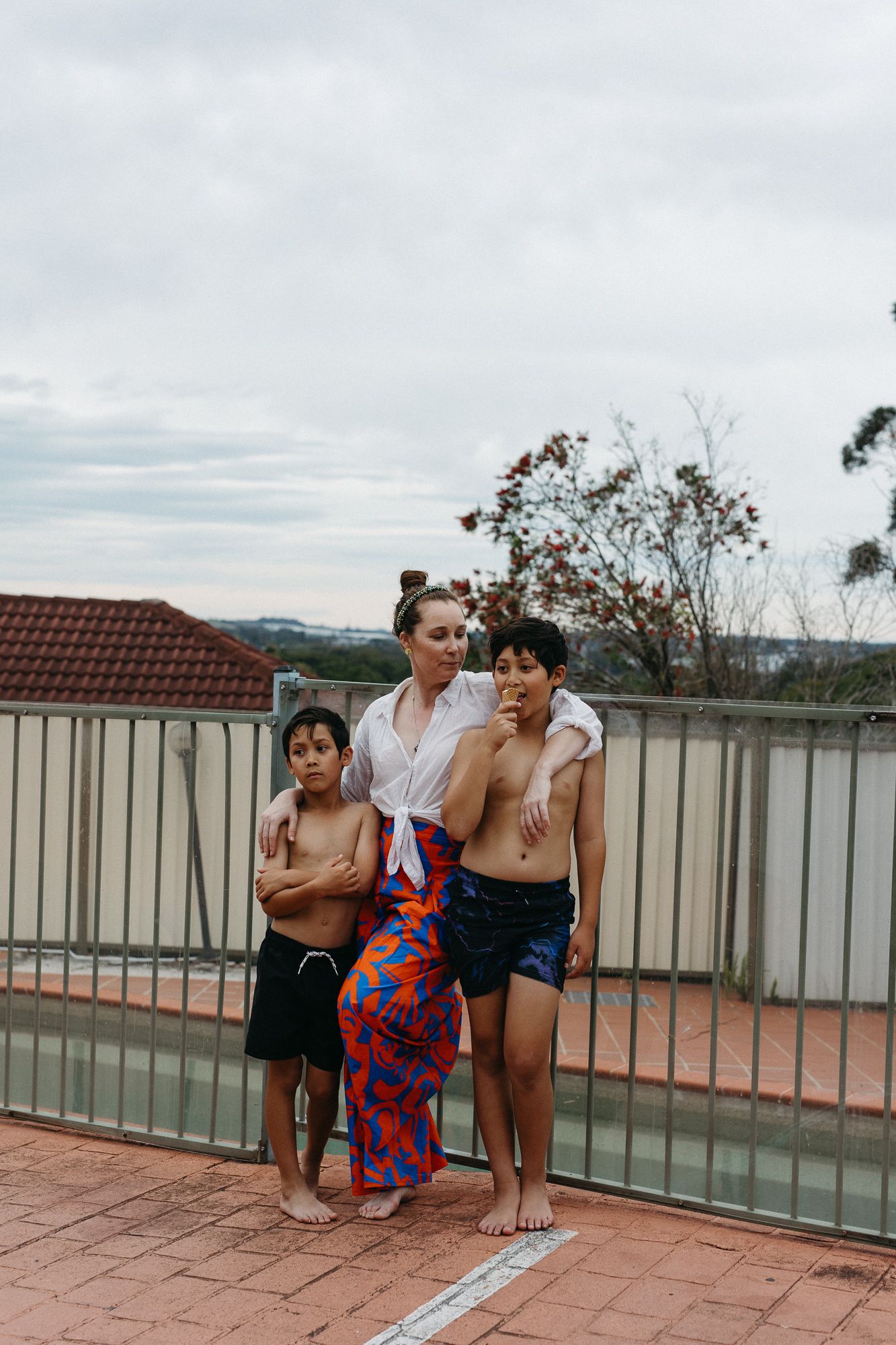 A woman in a white shirt and colorful skirt stands with two boys in swim trunks on a rooftop pool deck.