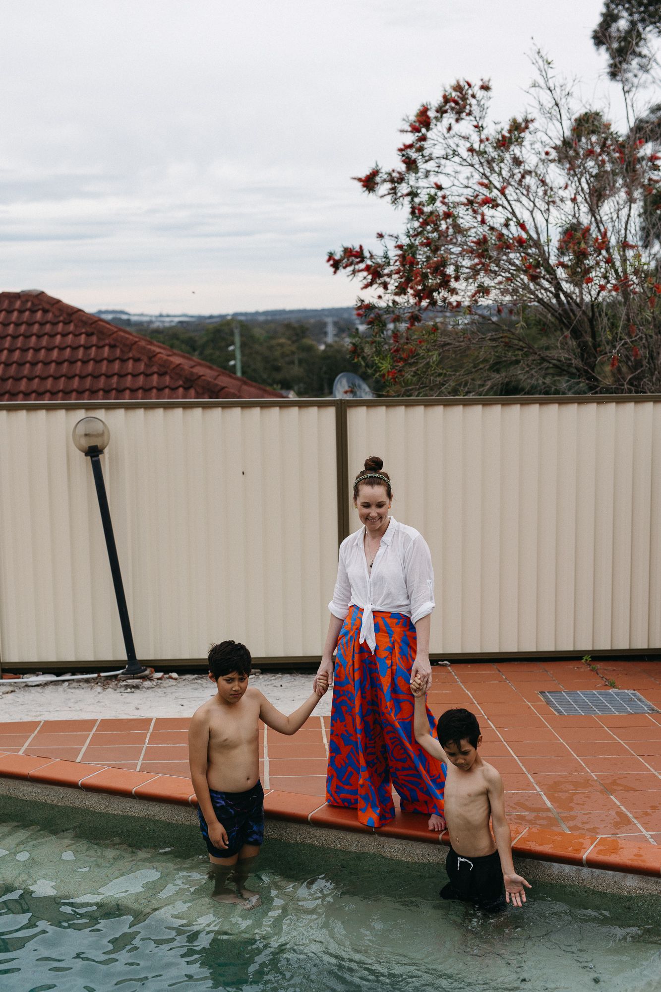 Woman in colorful maxi skirt holds hands with two boys standing in backyard swimming pool on cloudy day.