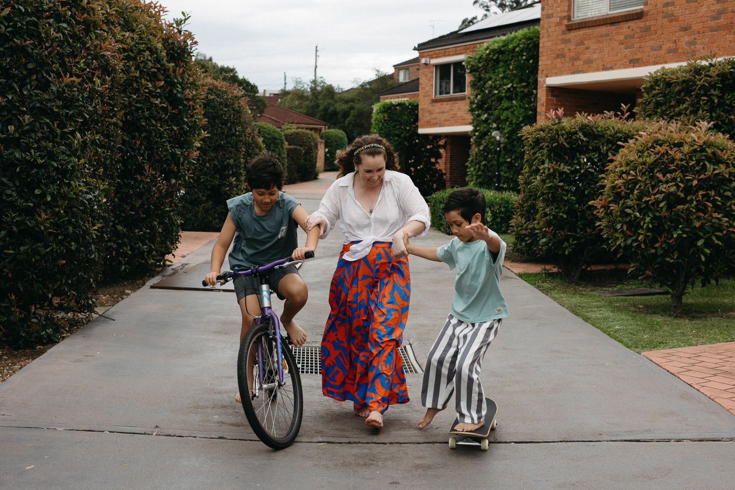 A mother helps her two kids ride a bike and skateboard on a suburban driveway.