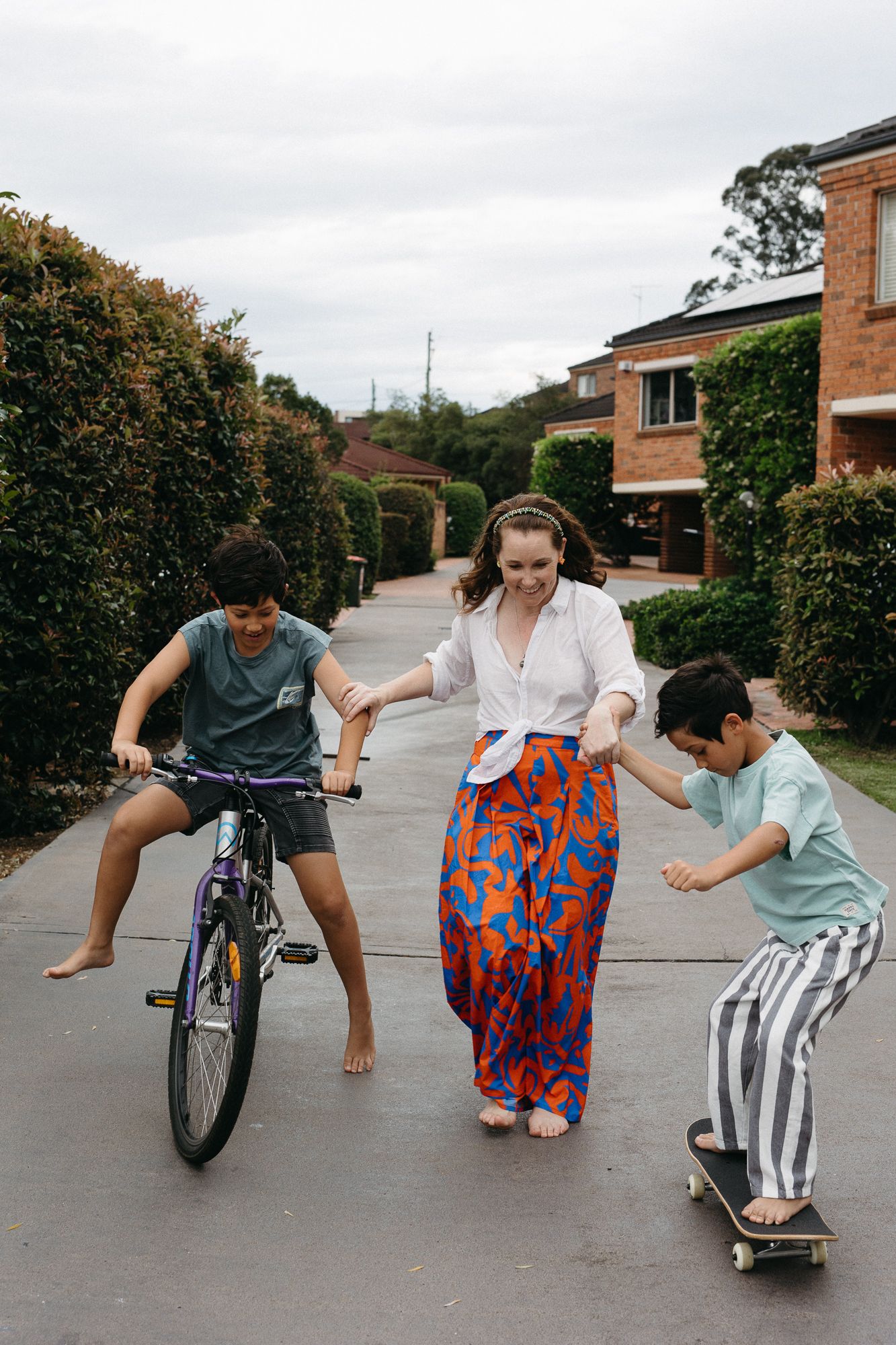 A woman helps two children play outside, one riding a bike and one skateboarding, on a suburban driveway.