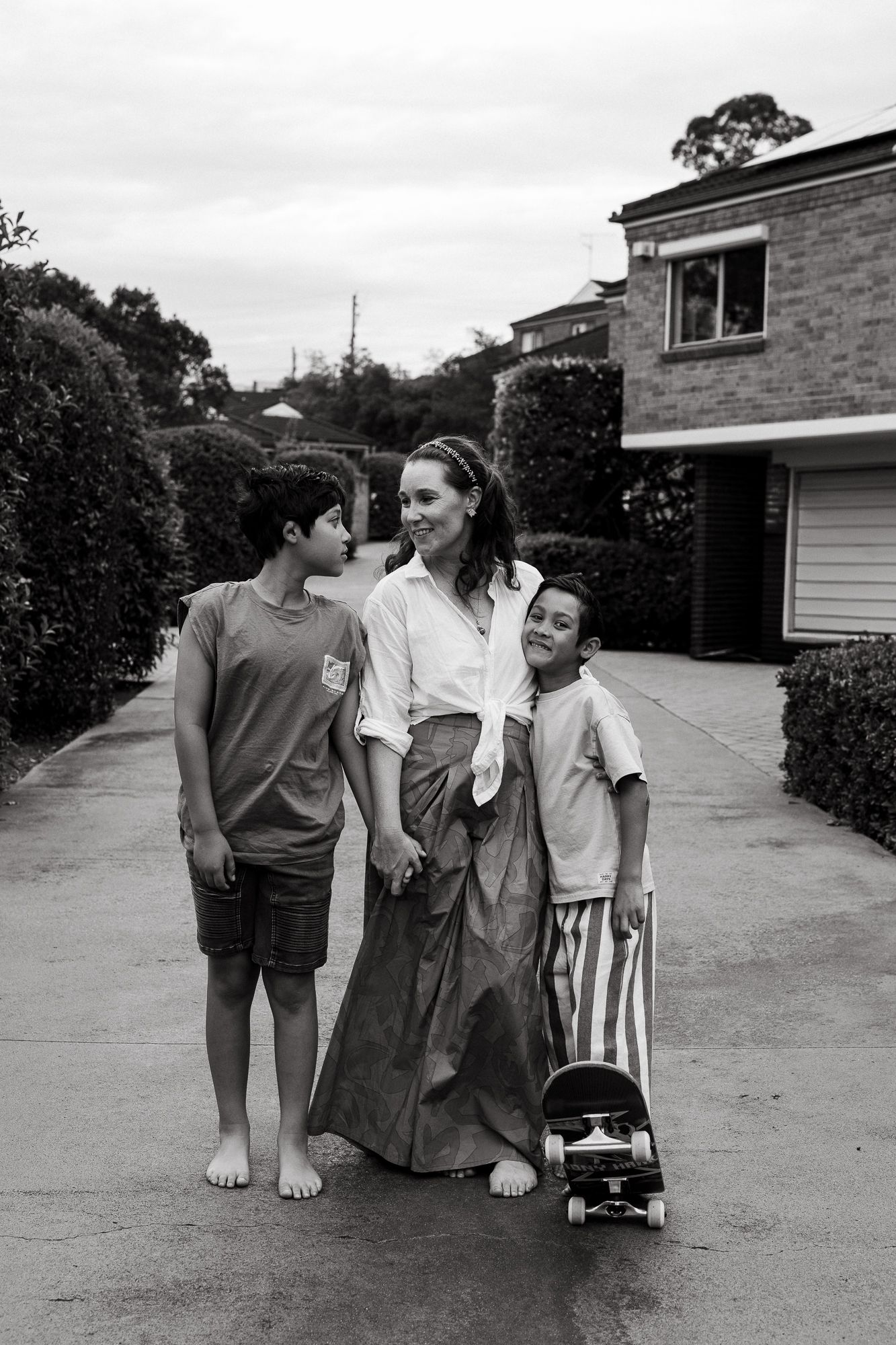 Black and white photo of a mother and her two young sons standing together on a suburban driveway, one boy holding a skateboard.