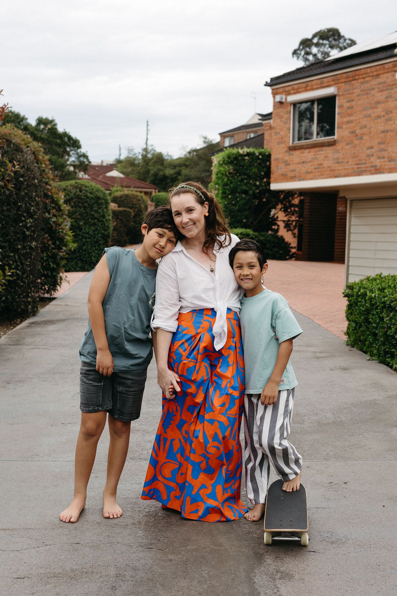 A woman in a colorful wrap skirt poses with two young boys on a suburban driveway, one holding a skateboard.