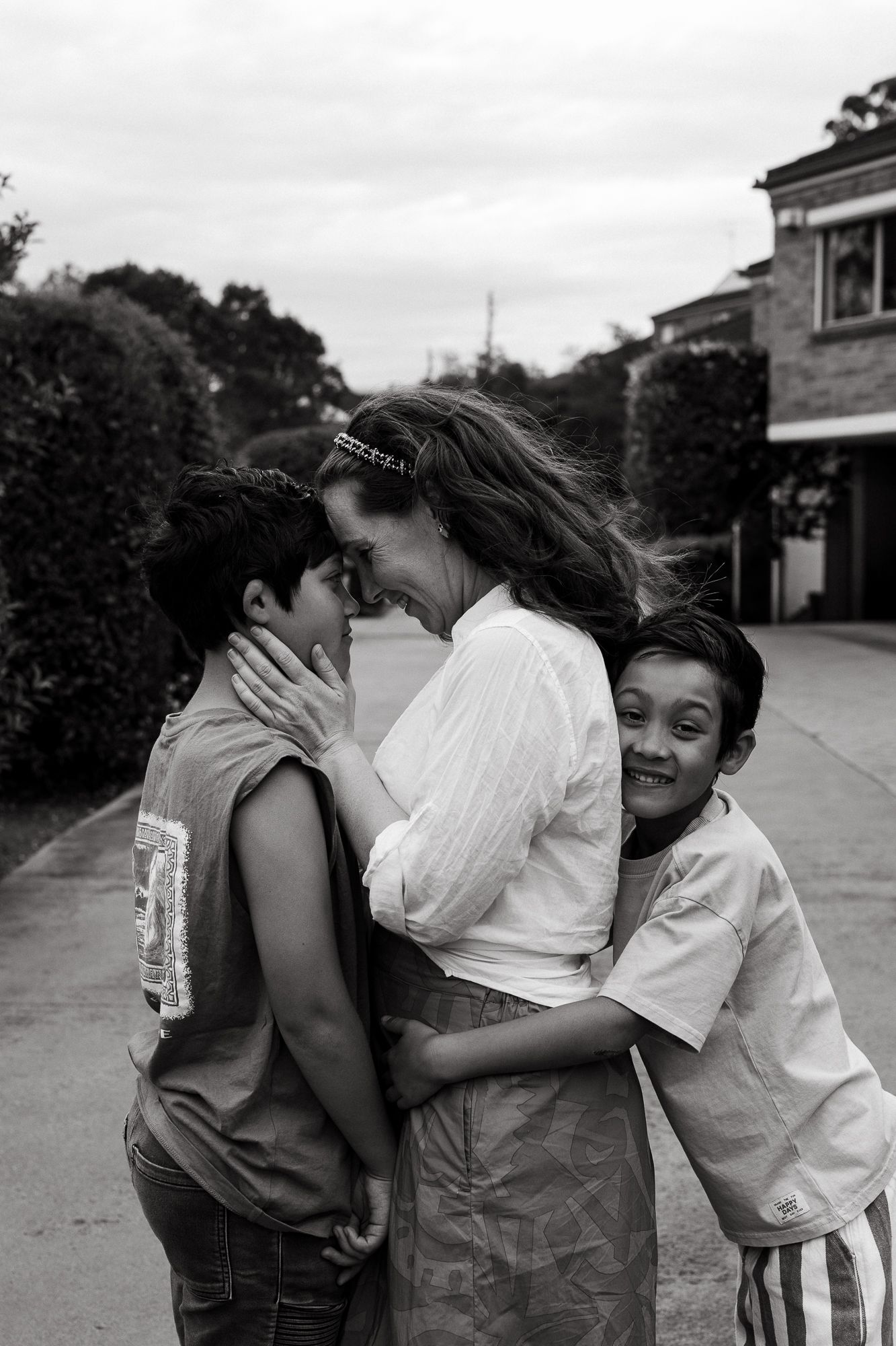 Black and white photo of a woman hugging two young boys on a suburban street, sharing a tender family moment.