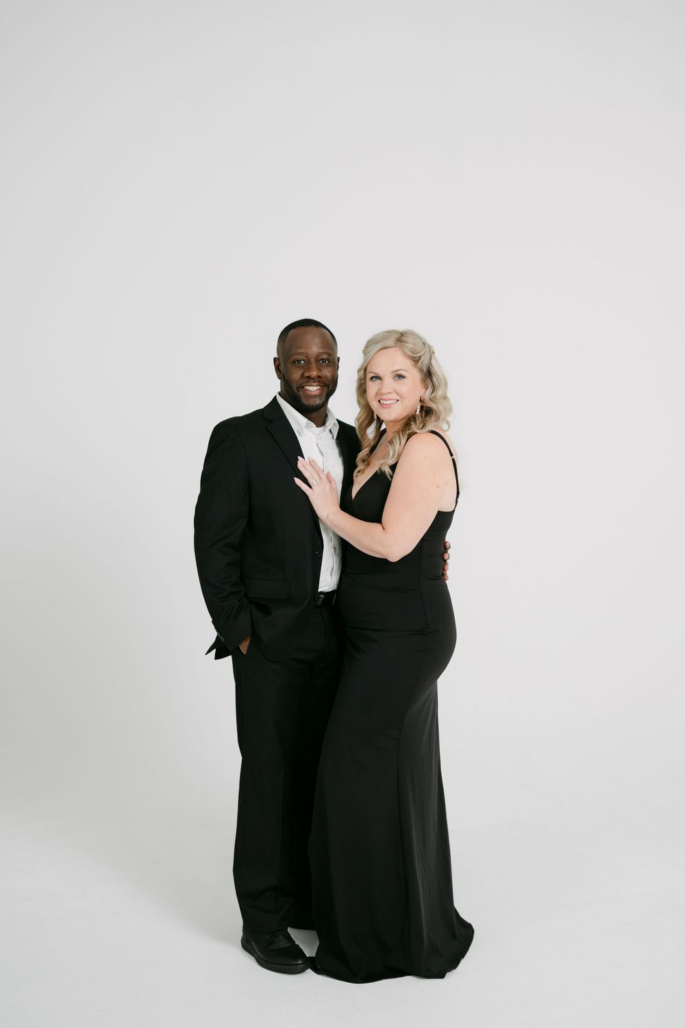 A smiling couple in formal black attire, a man in a suit and a woman in a long gown, pose together against a clean white studio backdrop.