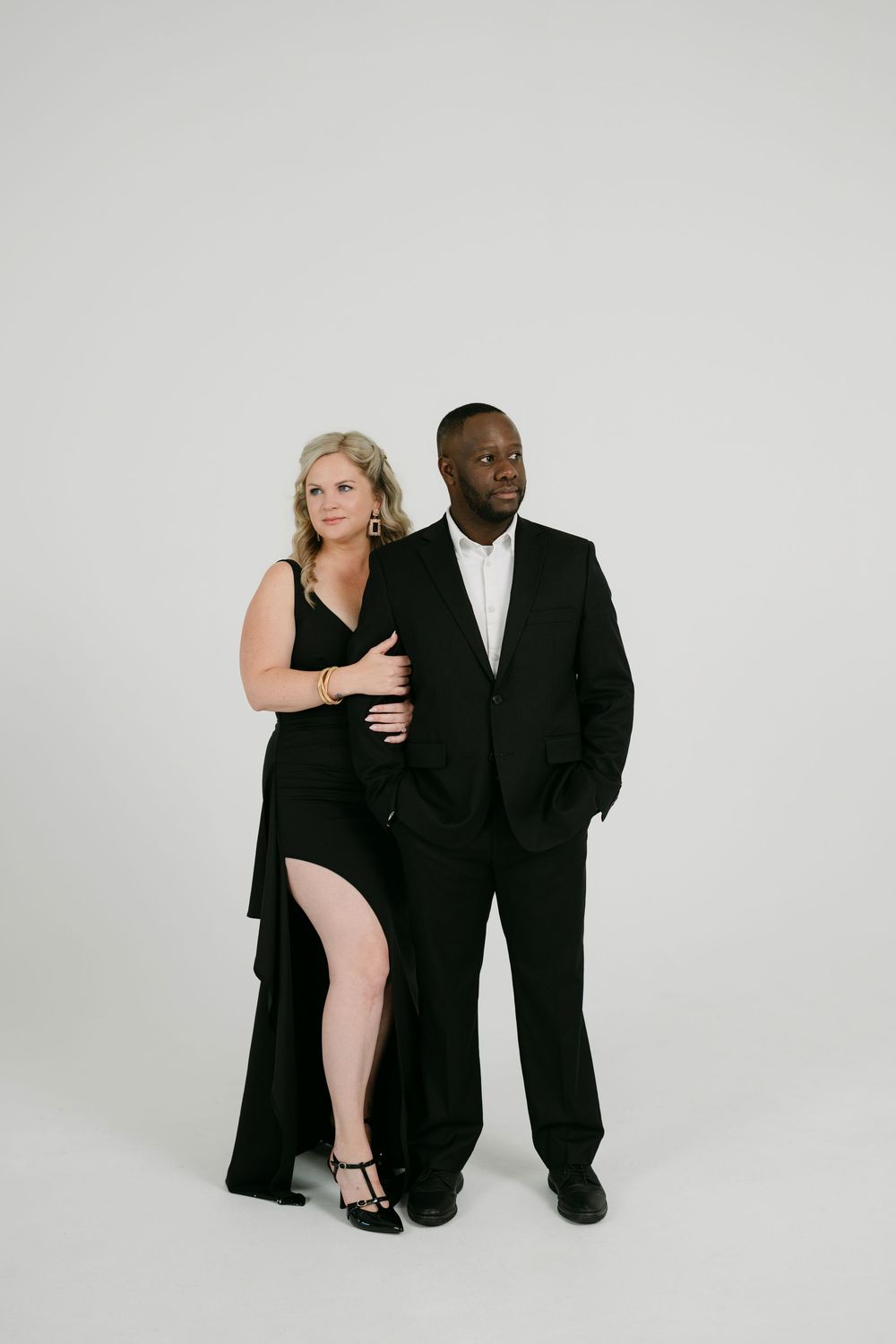 Couple in elegant black formal attire posing together against a plain white studio backdrop, the woman in a slit gown and the man in a suit.