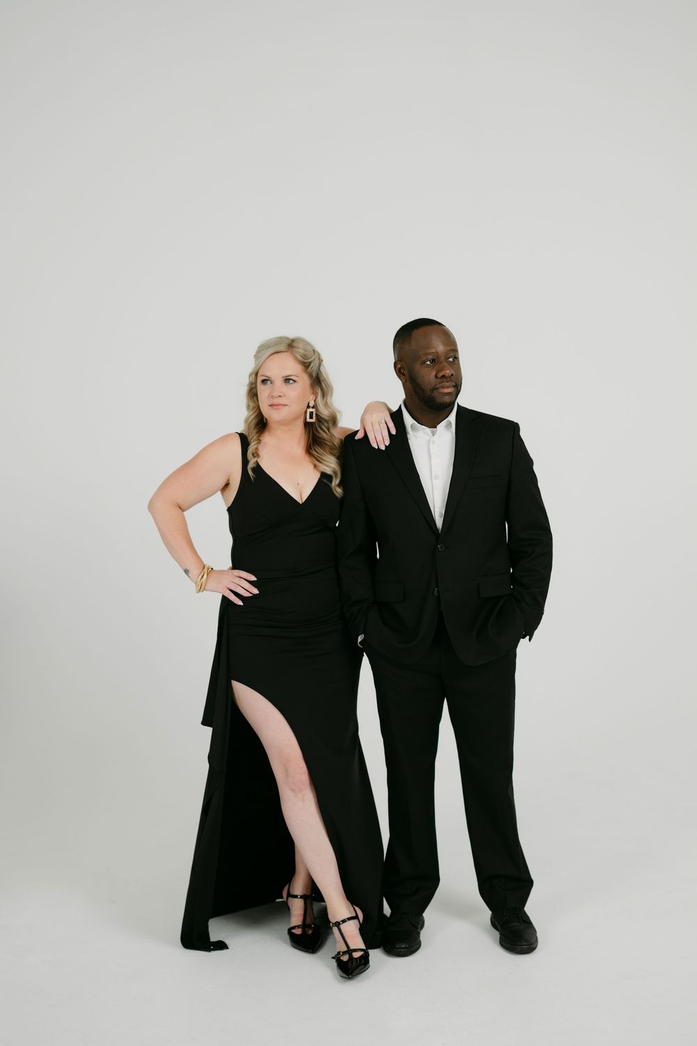 Woman in a black gown with a high leg slit leans on a man in a black suit and white shirt against a plain white studio backdrop.