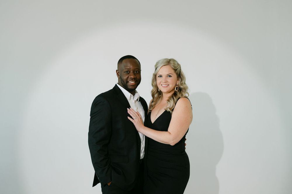 A smiling couple in elegant black formal attire poses closely together against a soft white studio backdrop.