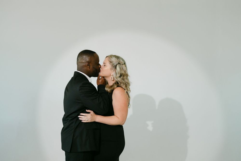 Couple in black outfits share an intimate kiss while embracing against a plain white wall with a soft spotlight and shadow.