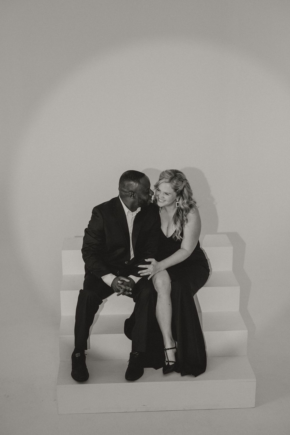 Black and white engagement portrait of a couple in formal black attire sitting close and laughing together on white studio steps.