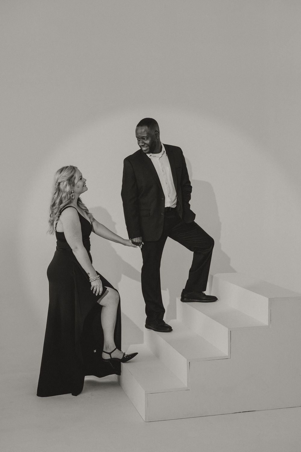 Black and white portrait of a woman in a long dress and a man in a suit holding hands on white studio stairs.