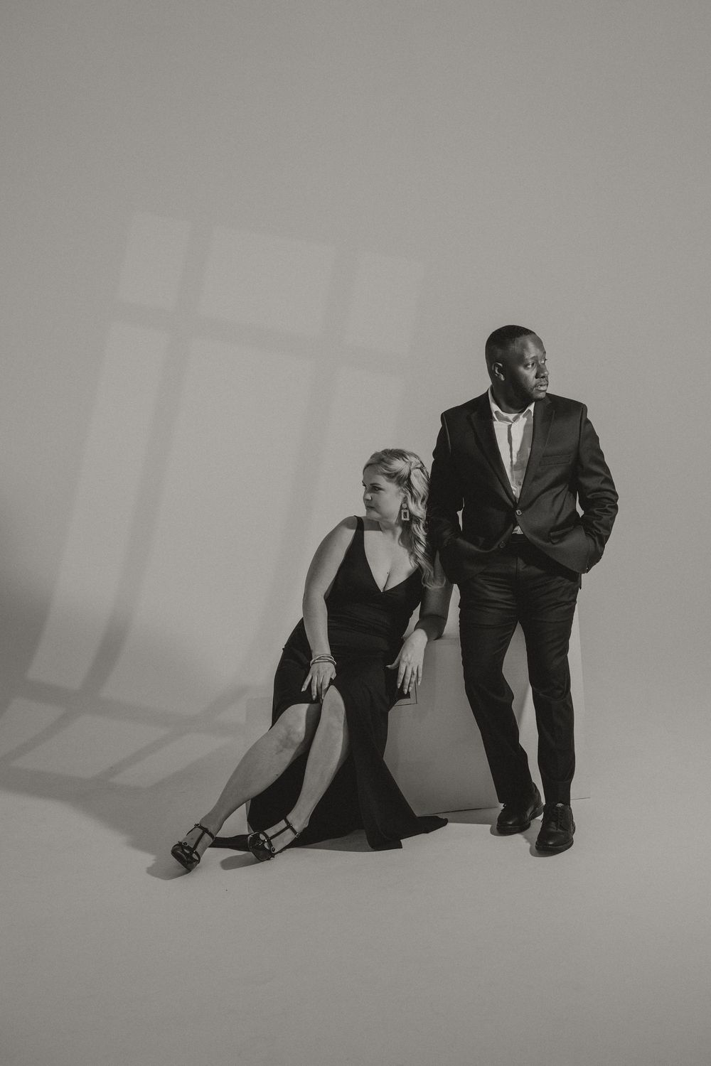 Black and white studio couple portrait of a woman in a black slit gown seated beside a man in a suit standing with hands in pockets.