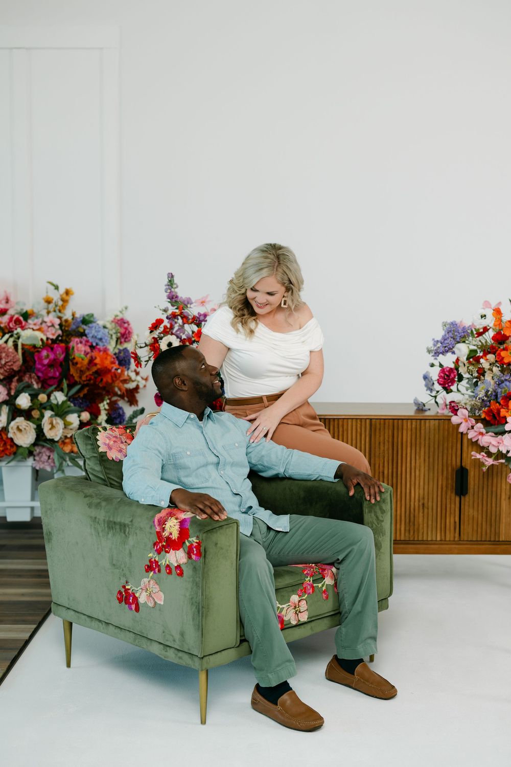 Smiling couple posing together on a green velvet armchair surrounded by colorful floral arrangements in a bright, modern studio.