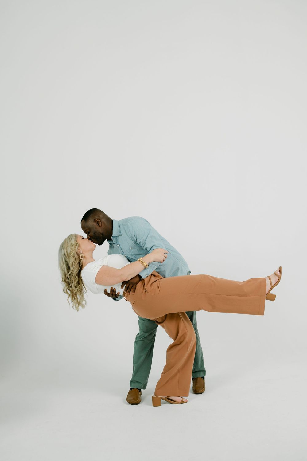 A couple shares a romantic dip kiss in a bright studio, she in a white top and rust pants, he in a blue shirt and green pants.